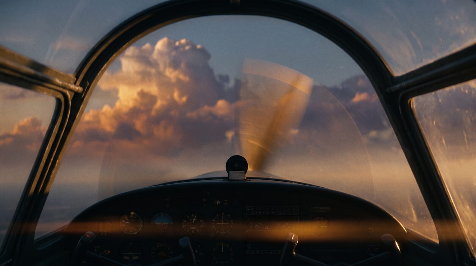 The view from a small aircraft cockpit at golden hour: propeller spinning, towering clouds ahead, the instrument panel below